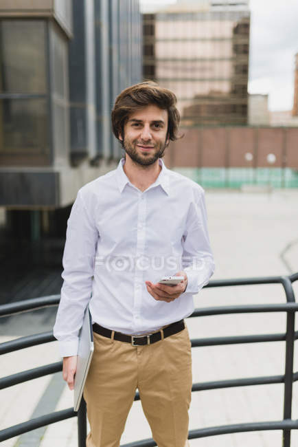 Retrato de hombre de negocios morena en camisa blanca posando con portátil y smartphone en las manos y mirando a la cámara en la escena urbana - foto de stock