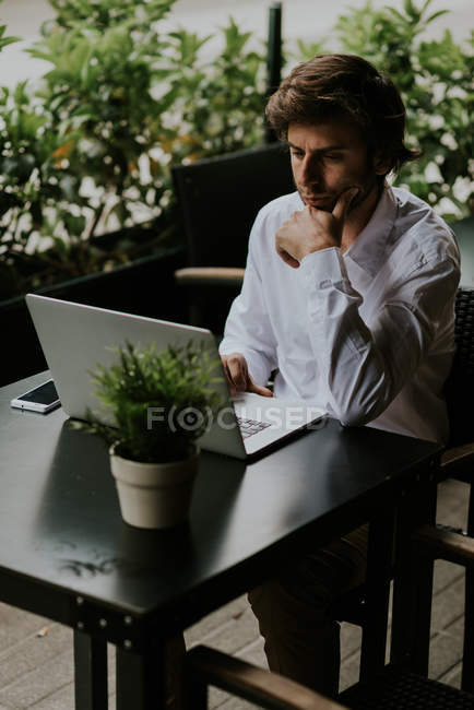 Portrait of confident businessman sitting at cafe terrace table with potted plant and using laptop — Stock Photo