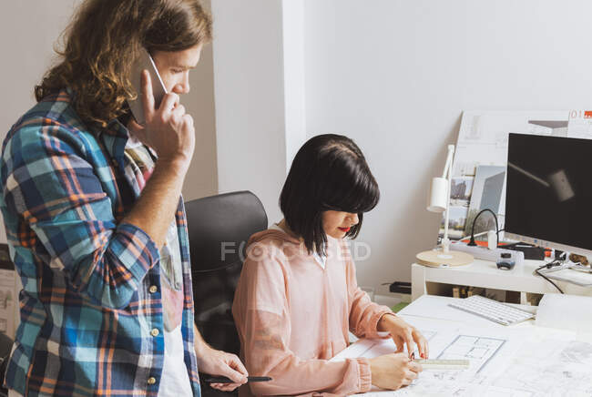 Side view of man talking phone and looking at woman sketching blueprint in the office — Stock Photo