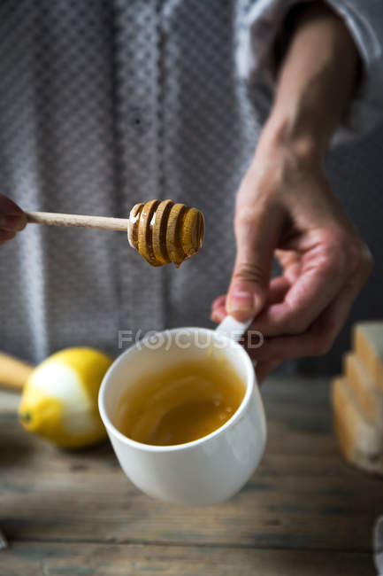 Couper les mains tenant une cuillère à miel au-dessus d'une tasse en céramique sur une table rurale en bois — Photo de stock