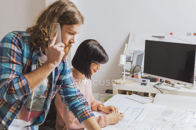 Portrait of man talking phone during  female colleague drawing blueprint in office. — Stock Photo