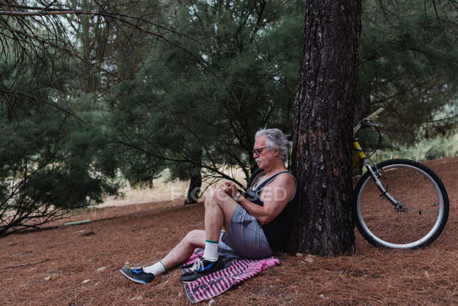 Vue latérale de l'homme âgé assis sur le sol avec livre dans les mains et appuyé sur l'arbre avec vélo garé — Photo de stock
