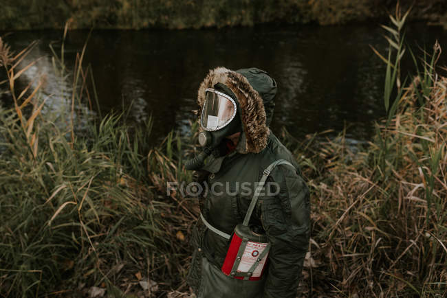 Vista de ángulo alto del hombre que usa abrigo con capucha y máscara de gas de pie en la orilla del lago - foto de stock