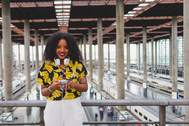 Retrato de una chica sonriente con auriculares en los hombros usando un teléfono inteligente y mirando la cámara en la estación de tren - foto de stock