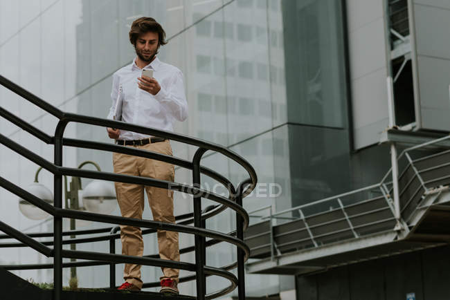 Портрет бизнесмена в белой рубашке, стоящего у лестницы, несущего ноутбук и использующего телефон над стеклянным фасадом бизнес-здания — стоковое фото