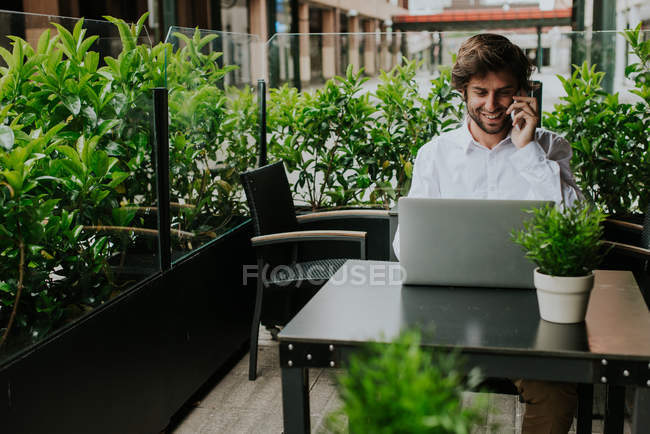 Усміхнений бізнесмен розмовляє по клітині, сидячи за столом кафе з ноутбуком — стокове фото