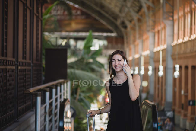 Bella ragazza sorridente che porta valigia su ruote e parla al telefono alla stazione ferroviaria — Foto stock