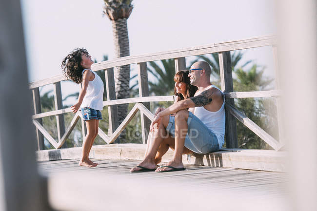 Los padres sentados junto a la barandilla y mirando a la hija saltando - foto de stock