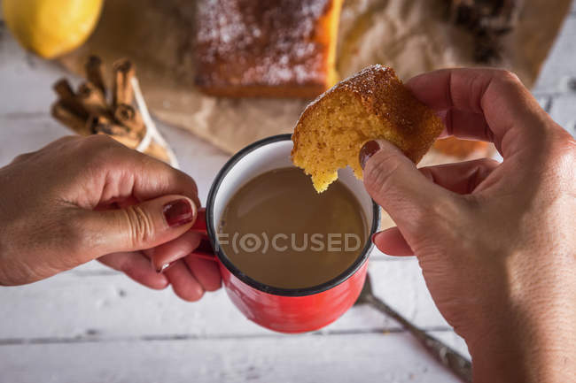 Sopra vista di mani femminili che tengono tazza di cioccolata calda e fetta di torta al limone sul tavolo della cucina rurale — Foto stock