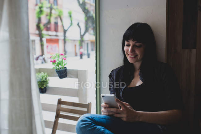 Mujer sentada cerca de la ventana y usando asmartphone - foto de stock