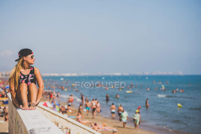 Elegante ragazza con berretto da baseball e occhiali da sole seduta sul parapetto sulla spiaggia contro la folla irriconoscibile di persone — Foto stock