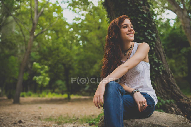 Портрет девушки с длинными вьющимися рыжими волосами, сидящей на камне в лесу и смотрящей в сторону — стоковое фото