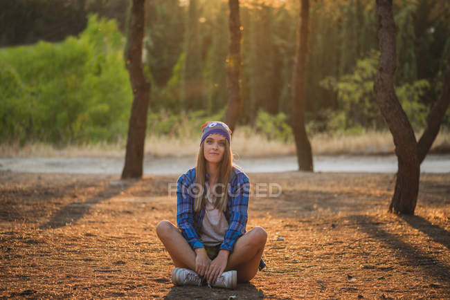 Ritratto di donna con divertente cappello di lana seduta a terra nella foresta e che guarda la macchina fotografica — Foto stock