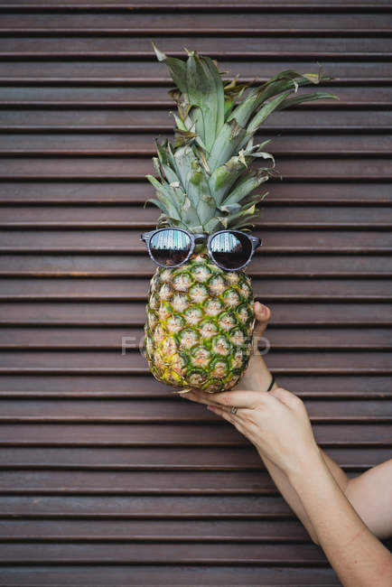 Weibliche Hand hält lustige Ananas mit Sonnenbrille über braunen Rollläden — Stockfoto