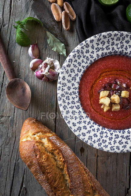 Томатный суп в узорной тарелке на деревянном столе с хлебом и ингредиентами — стоковое фото