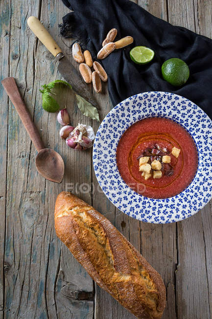 Sopa de tomate em prato estampado em mesa de madeira com pão e ingredientes — Fotografia de Stock