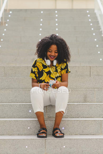 Retrato de mulher alegre com fones de ouvido em torno do pescoço usando smartphone e olhando para longe enquanto sentado em escadas — Fotografia de Stock