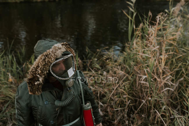 Vista de alto ângulo do homem vestindo casaco com capuz e máscara de gás em pé na margem do lago e olhando para longe — Fotografia de Stock