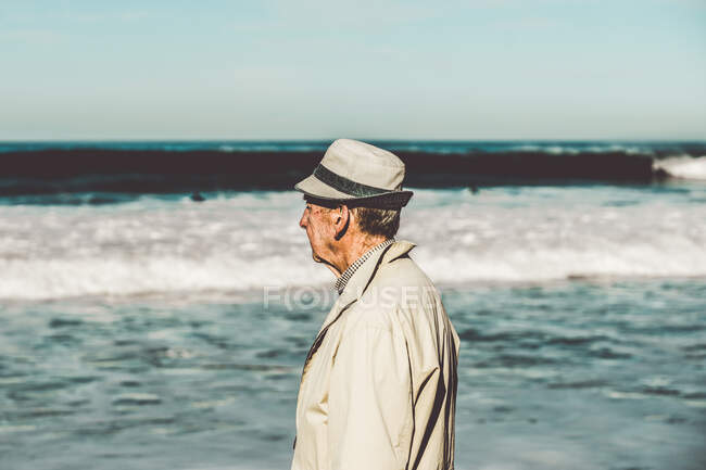 Vista lateral do homem adulto sênior em um chapéu e casaco assistindo oceano ondulado à luz do sol — Fotografia de Stock