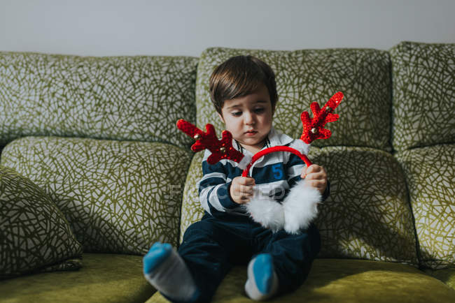 Porträt eines Kleinkindes, das auf einem Sofa sitzt und Ohrenschützer aus Spielzeugfell mit Geweih hält — Stockfoto