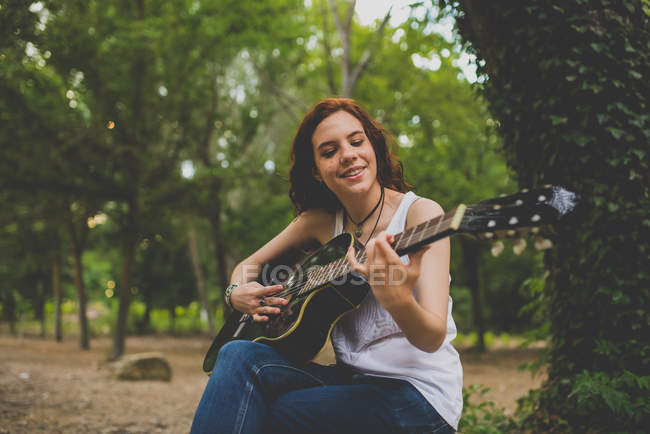 Retrato de niña pecosa sonriente sentada en piedra y tocando la guitarra en el bosque - foto de stock