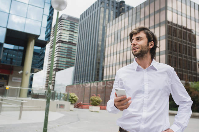 Портрет брюнетки бізнесмен тримає смартфон в руці і дивиться далеко на міську сцену в центрі міста — стокове фото