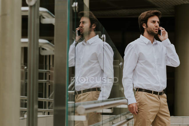 Recinzione in vetro che riflette l'uomo d'affari bruna in camicia bianca che parla sullo smartphone sulla scena urbana — Foto stock