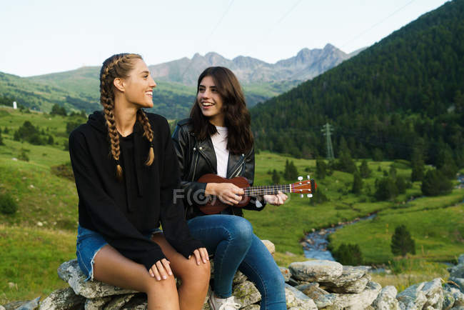 Cheerful women sitting on rocks at meadow and playing ukulele — Stock Photo
