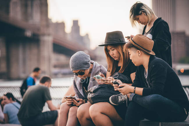 Um pequeno grupo de amigos todos usando smartphones na cidade contra rio e ponte — Fotografia de Stock
