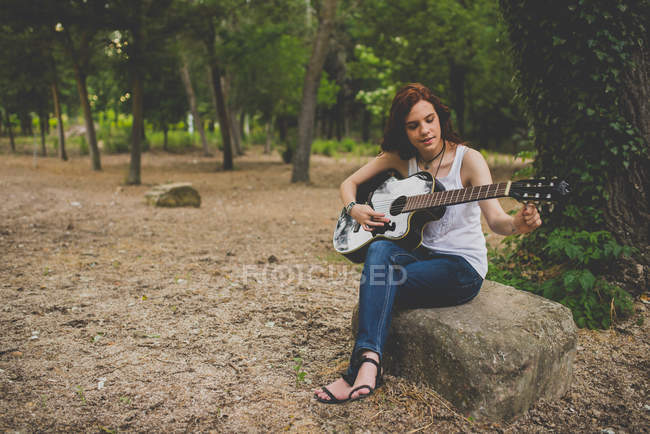 Portrait of smiling freckled girl sitting on stone and playing guitar and — Stock Photo
