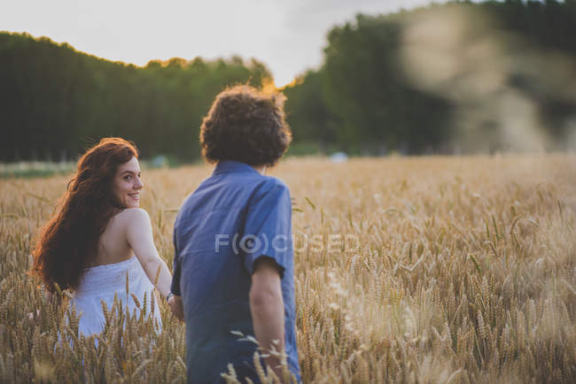 Visão traseira da menina de cabelos vermelhos olhando sobre o ombro e levando namorado para campo de centeio — Fotografia de Stock