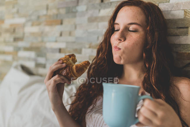 Girl eating croissant with cup of coffee in bed — Stock Photo