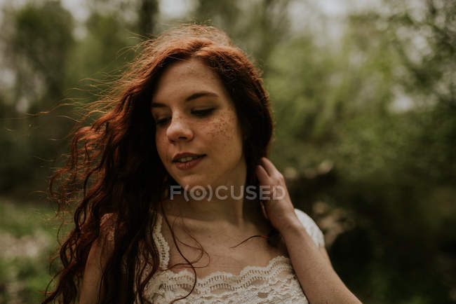 Чувственная рыжая девушка в белом платье трогает волосы и смотрит вниз — стоковое фото