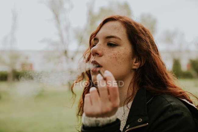 Рыжая девушка с веснушками курит сигарету на природе — стоковое фото