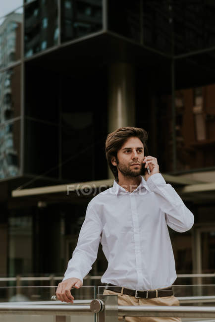 Retrato de empresario confiado en camisa blanca hablando por teléfono inteligente y mirando a un lado en la escena urbana - foto de stock