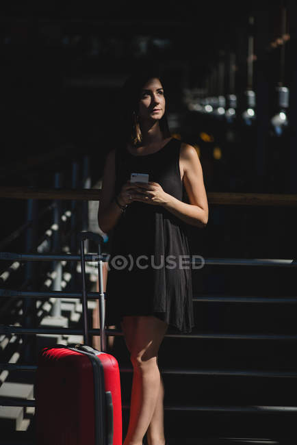 Portrait of brunette woman with red suitcase standing in sunlight and holding smartphone in hands — Stock Photo