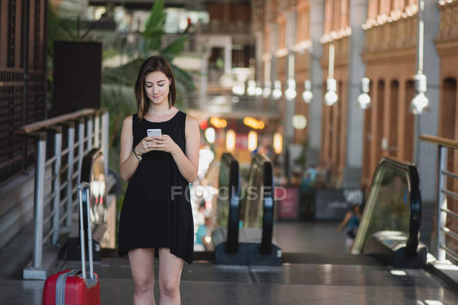 Retrato de una joven con una maleta roja usando un teléfono inteligente y de pie cerca de una escalera mecánica en un gran salón - foto de stock