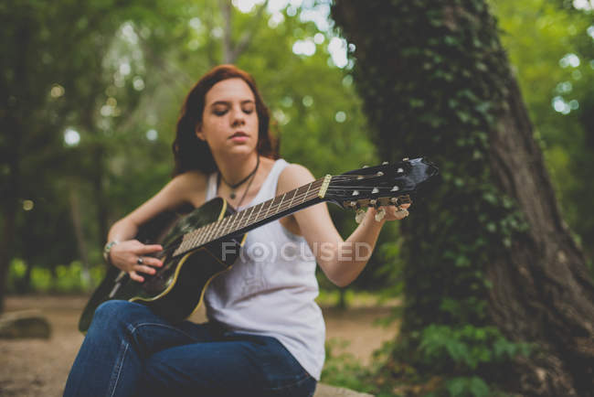 Portrait of freckled girl with long curly hair sitting at woods and tuning guitar — Stock Photo