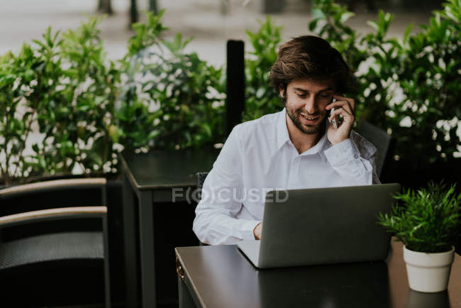 Porträt eines lächelnden Geschäftsmannes, der am Tisch sitzt und über sein Smartphone spricht, während er Laptop auf der Caféterrasse benutzt — Stockfoto