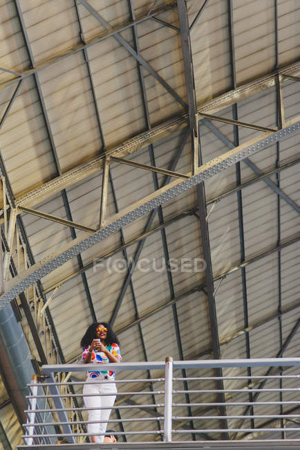 Vista a basso angolo di ragazza con smartphone in mano appoggiata su ringhiera balcone sotto la costruzione del tetto in ferro . — Foto stock