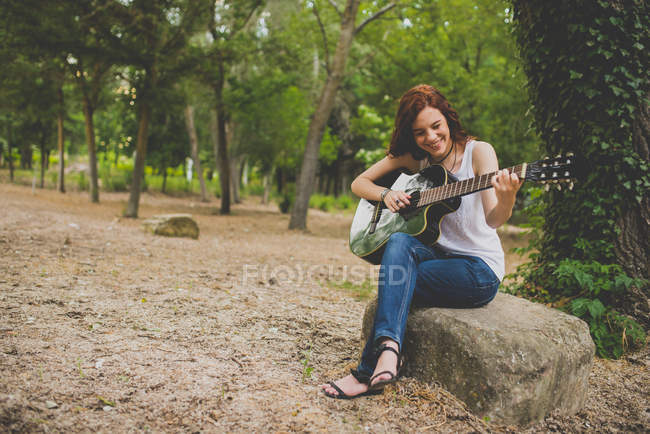 Retrato de niña pecosa riendo sentada en piedra y tocando la guitarra en el bosque - foto de stock