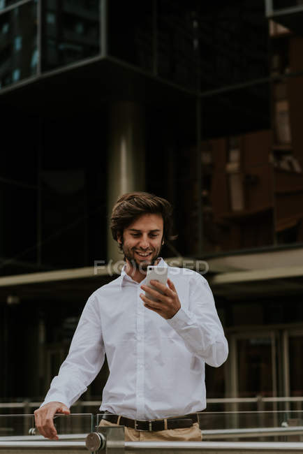 Retrato del hombre de negocios sonriente en camisa blanca mirando el teléfono inteligente en la mano en la escena urbana - foto de stock
