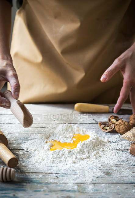 Close up view of female hands with scoop over pile of flour with smashed egg on rustic wooden table — Stock Photo
