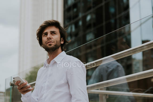 Retrato de empresario confiado en camisa blanca sosteniendo teléfono inteligente y mirando a un lado en la escena urbana - foto de stock