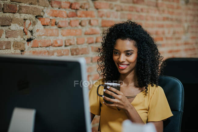 Усміхнена жінка тримає чашку сидячи за комп'ютером. Горизонтальний постріл у приміщенні . — стокове фото