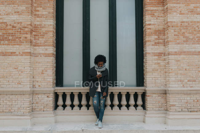 Retrato de una chica con ropa casual apoyada en la pared y usando un smartphone en la escena callejera - foto de stock