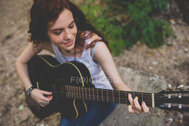 High angle portrait of smiling freckled girl sitting on rock and playing guitar — Stock Photo
