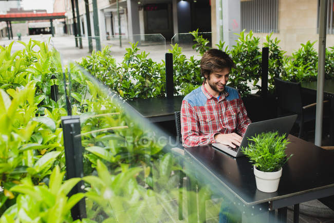 High angle portrait of bearded man in checkered shirt sitting at cafe terrace table with potted plant and using laptop over urban scene on background — Stock Photo