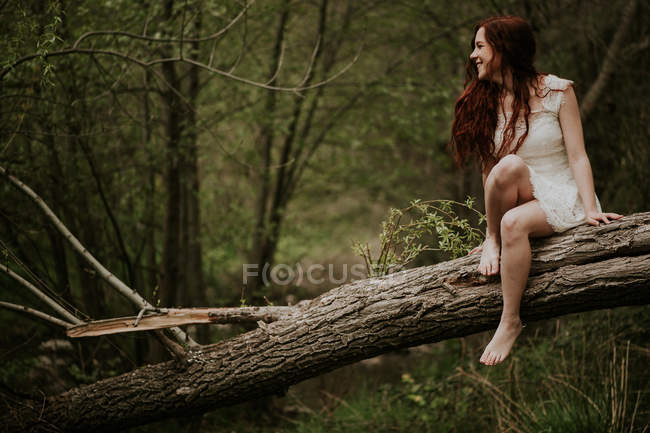 Chica alegre en vestido blanco sentado descalzo en el árbol caído - foto de stock