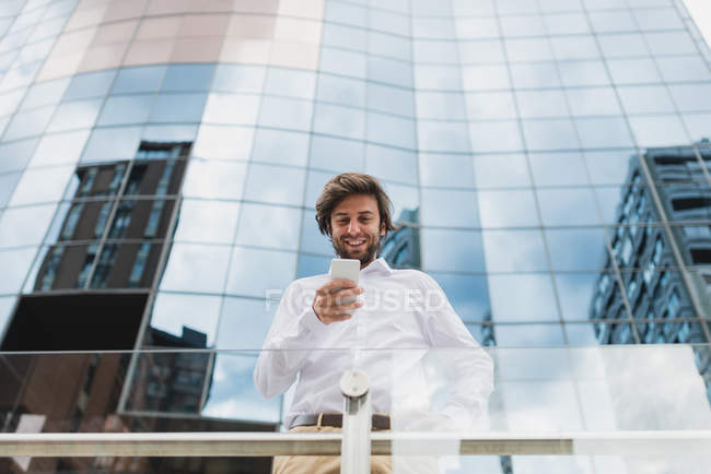 Портрет усміхненого бізнесмена в білій сорочці з використанням телефону над фасадом бізнес-будівель — стокове фото